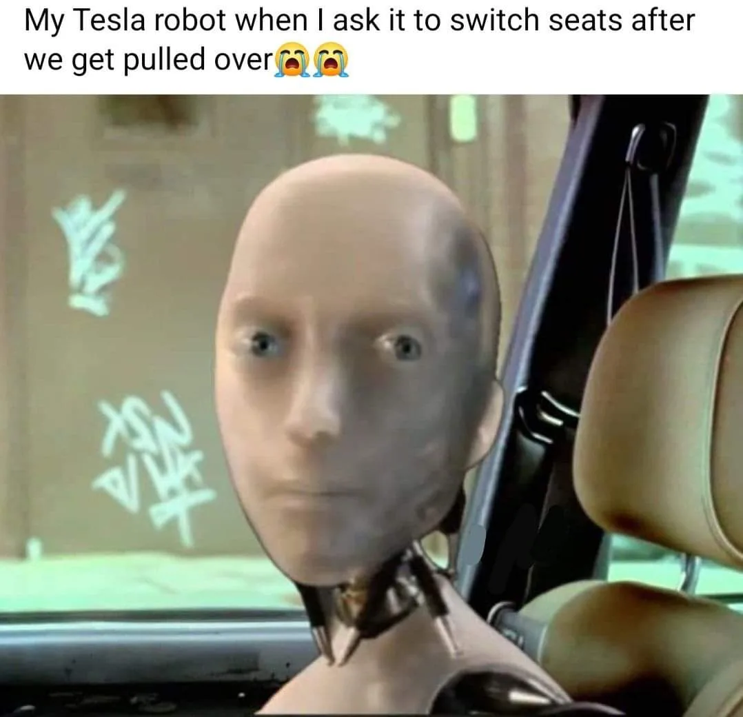 A bald, humanoid robot with a human-like face sits in a car seat, looking shocked and distressed. The overlaid text reads…