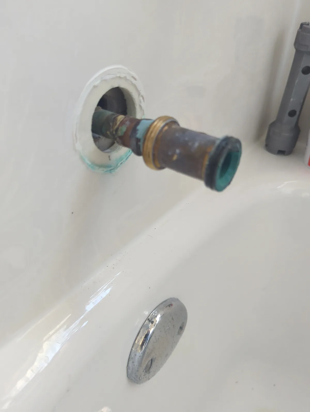 A close-up photo shows a rusty, corroded metal drain pipe protruding from a hole in the side of a white bathtub. The pipe…