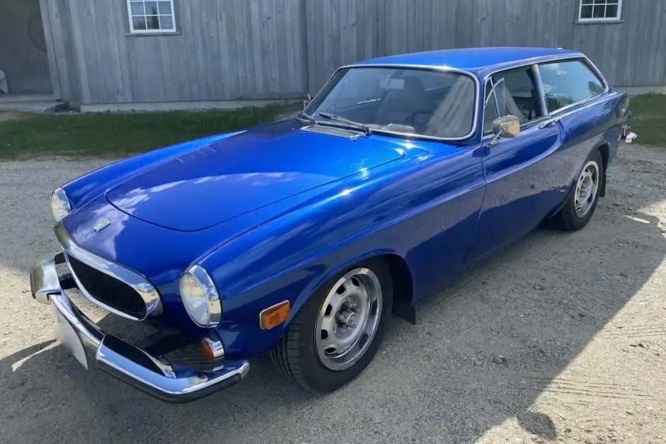 A shiny blue Volvo P1800 ES station wagon is parked on gravel in front of a wooden building. The car has chrome bumpers and…