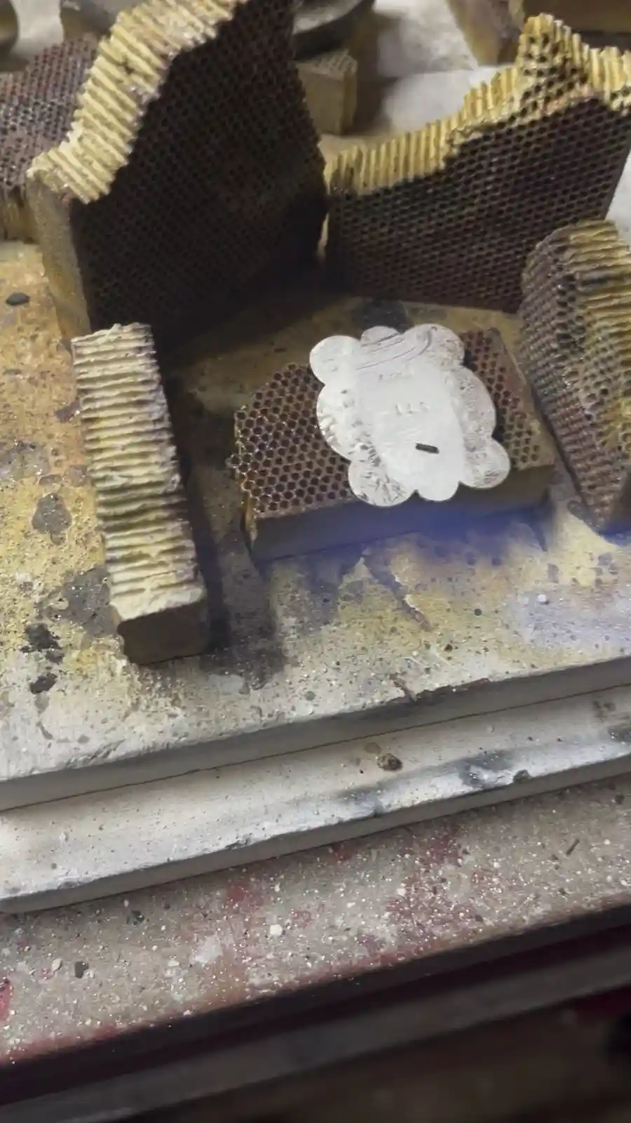A close-up of a small, silver, cloud-shaped metal piece resting on a honeycomb-patterned brick in a workshop. Surrounding it…