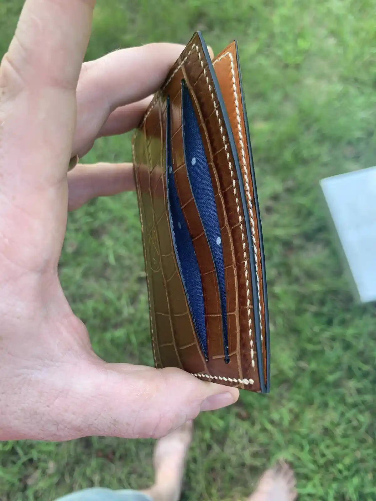 A hand holds a brown, crocodile-textured leather wallet open to reveal an empty card slot with only a blue fabric insert…