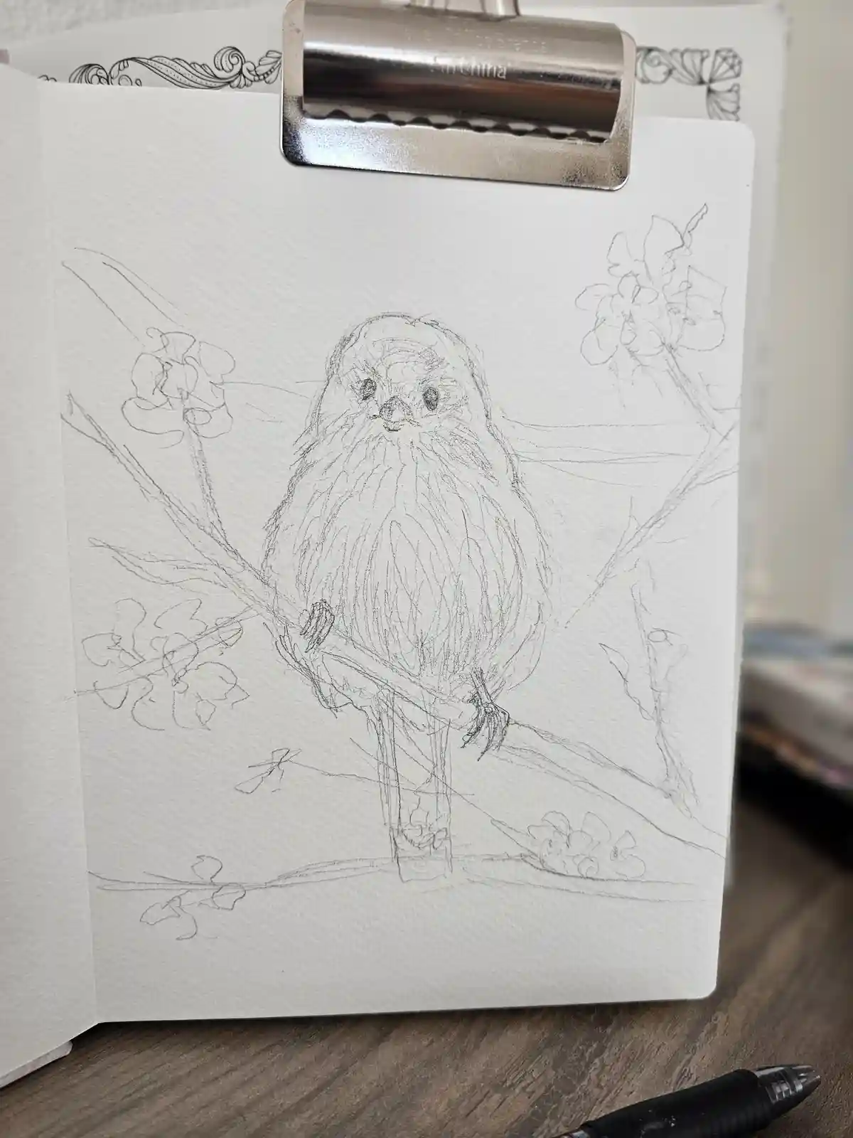 A pencil sketch of a bird perched on a branch with flowers, held in a clipboard on a wooden surface, with a black pen…