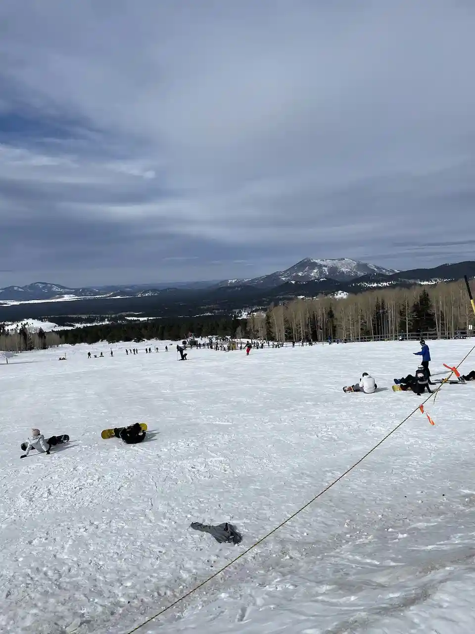 A wide shot of a snowy ski slope with numerous people scattered across the terrain, many sitting or lying down on the snow…
