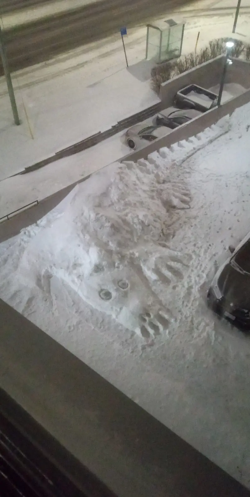 An aerial nighttime photo shows a snow-covered street with a large, crudely shaped snow figure resembling a monster with…