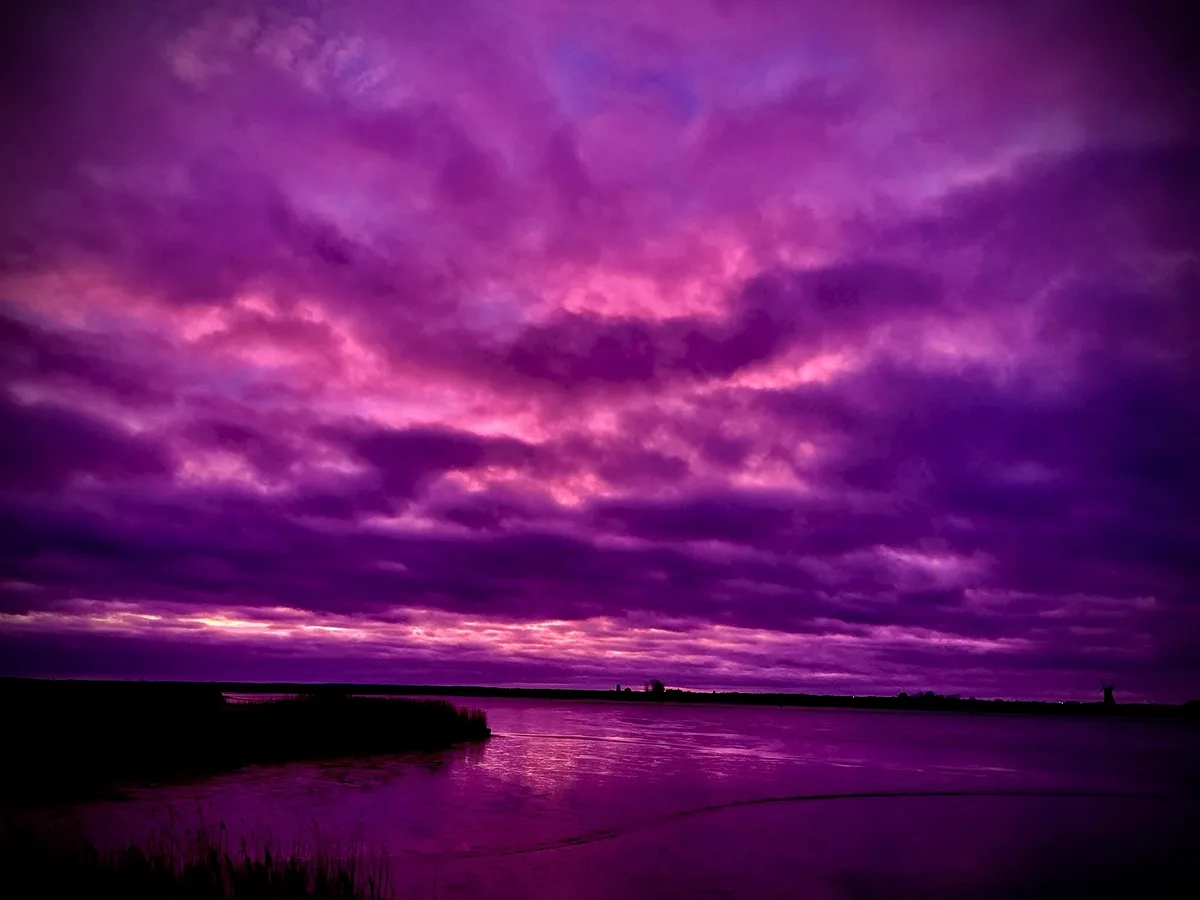 A dramatic landscape photo shows a body of water reflecting a sky filled with intense purple and pink clouds, with…