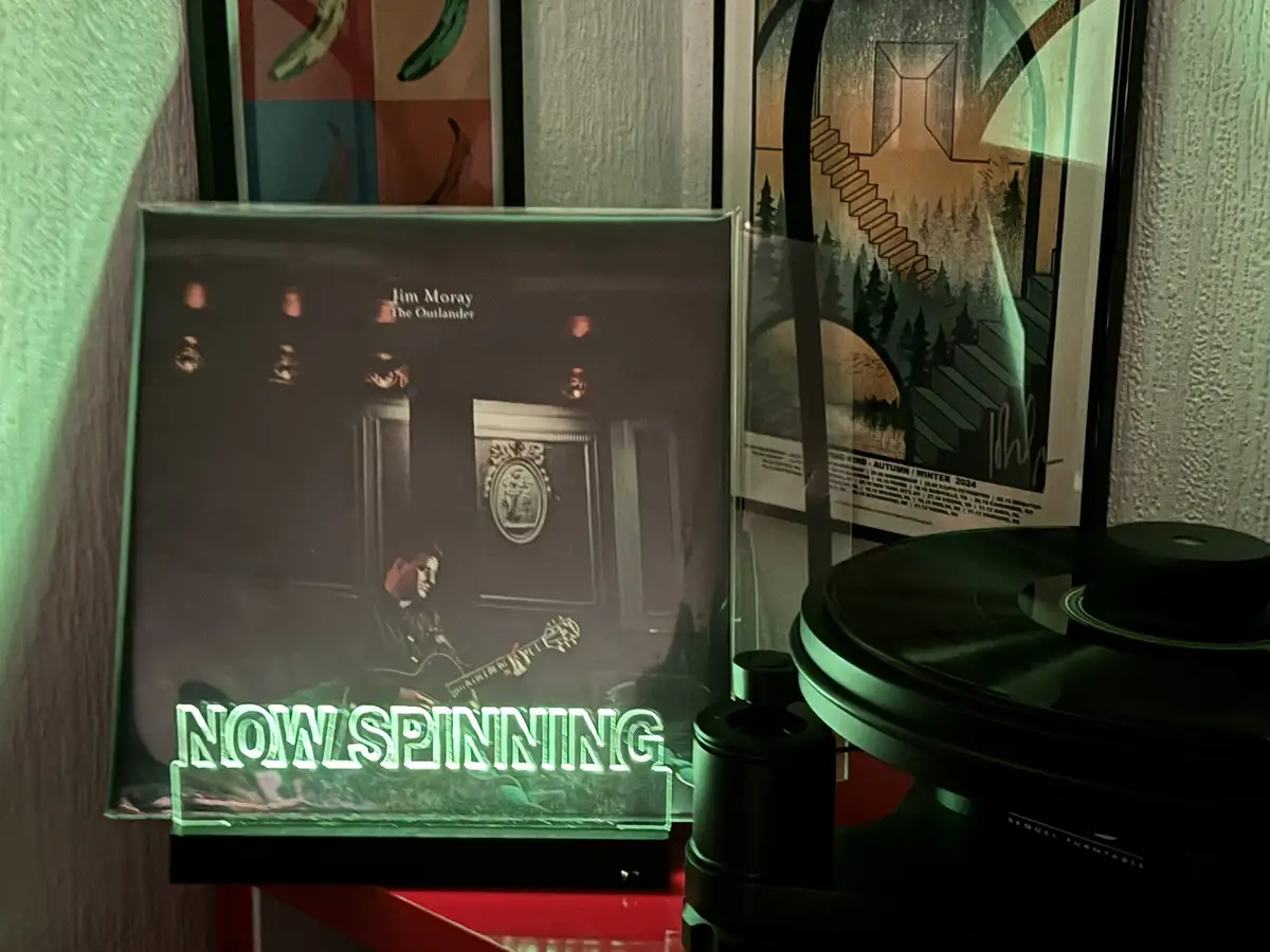 A close-up photo shows a vinyl record player with a glowing green 'NOW SPINNING' sign in front of it. The album cover for…