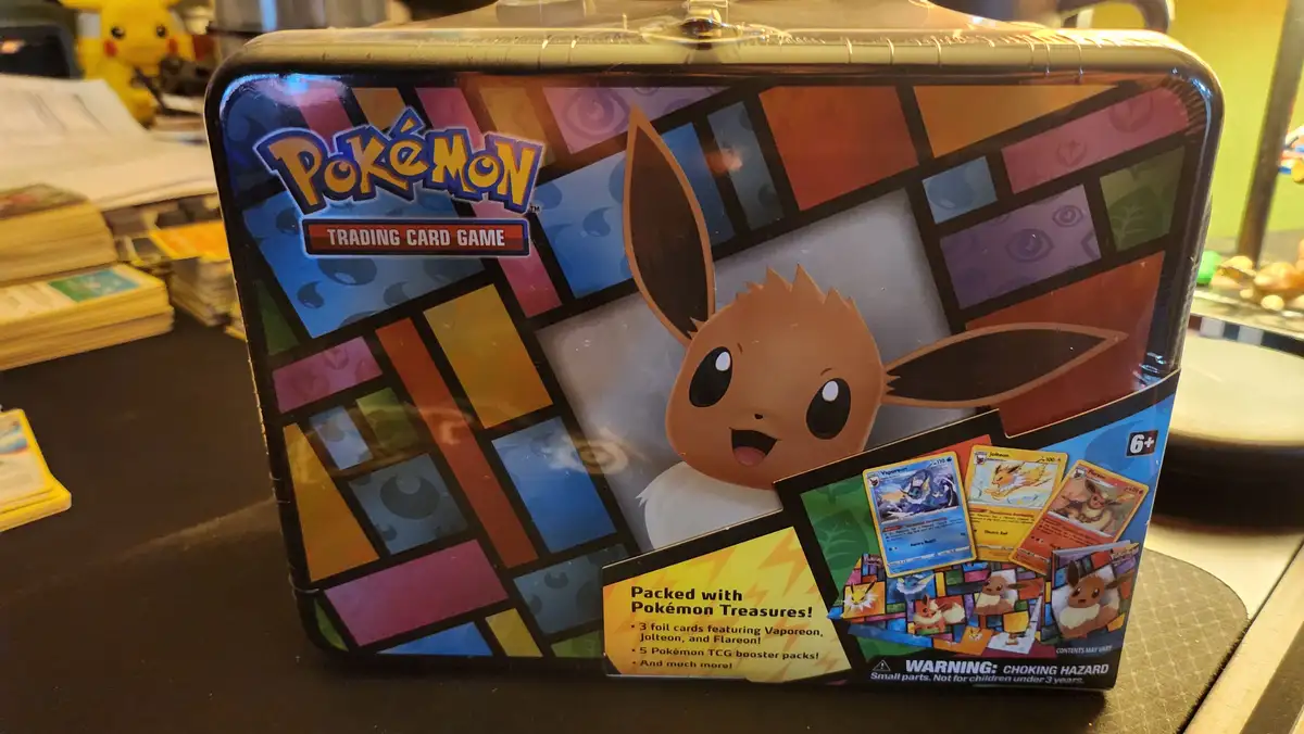 A Pokémon Trading Card Game tin featuring Eevee on a stained-glass style background, with text listing included items like…