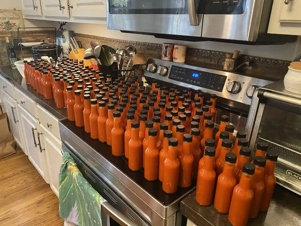 A kitchen counter and stovetop are covered with hundreds of identical small glass bottles filled with orange-red hot sauce…