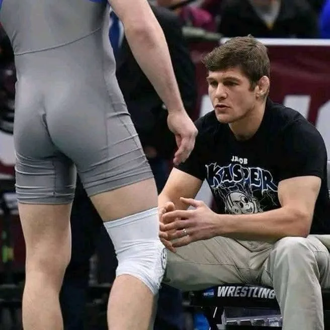 A wrestler in grey singlet and white knee brace stands with his back to the camera, while seated Jacob Kasper, wearing a…
