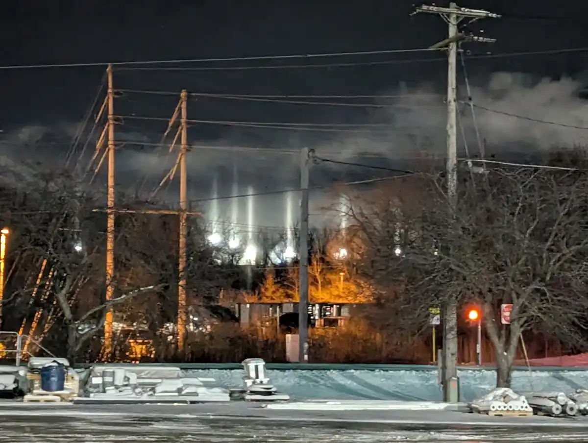 A nighttime photo of a snowy, industrial-looking area with utility poles, bare trees, and a distant lit building. In the…