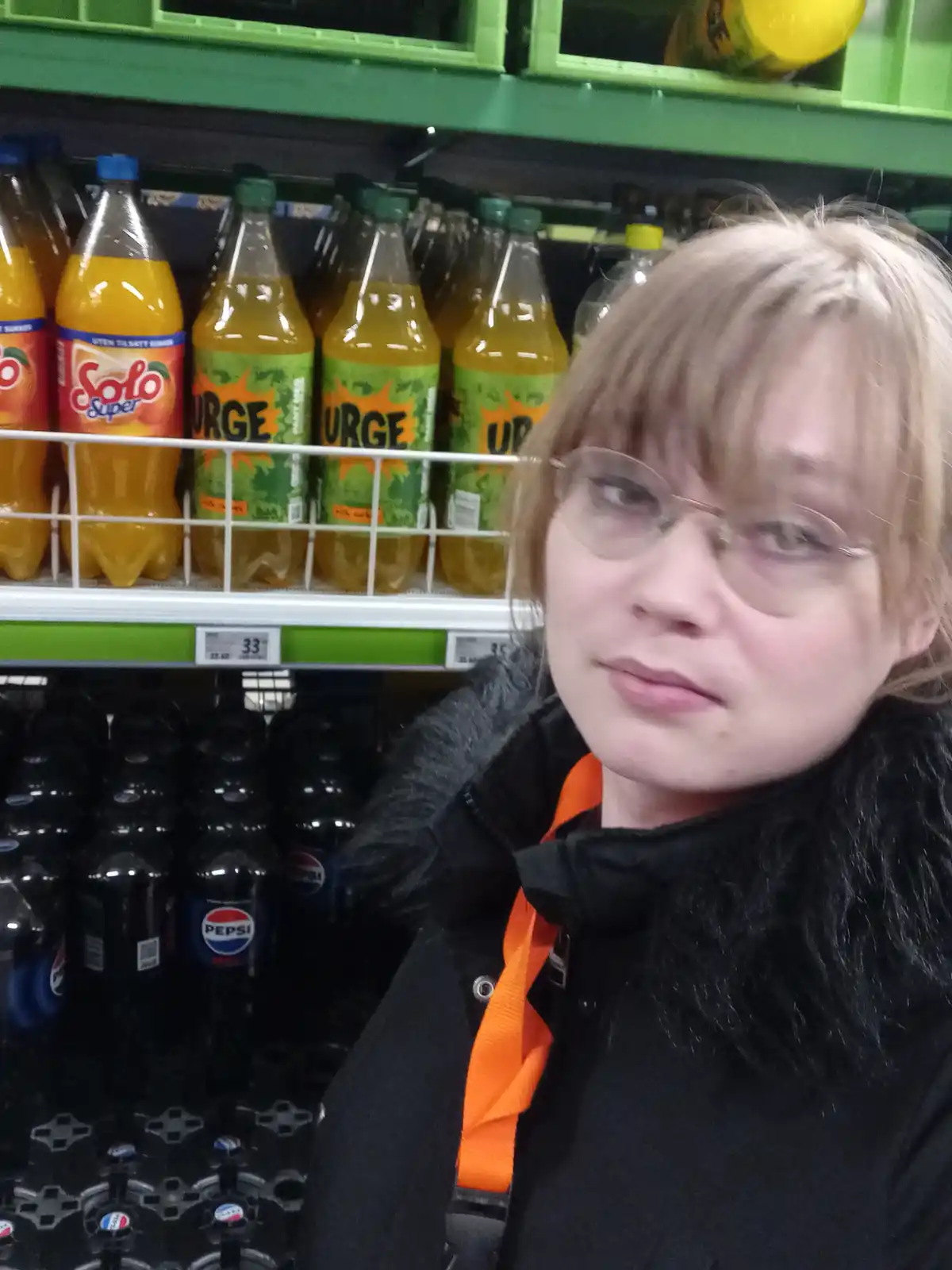 A person with glasses and a black jacket stands in a grocery store aisle, looking at the camera with a skeptical or…