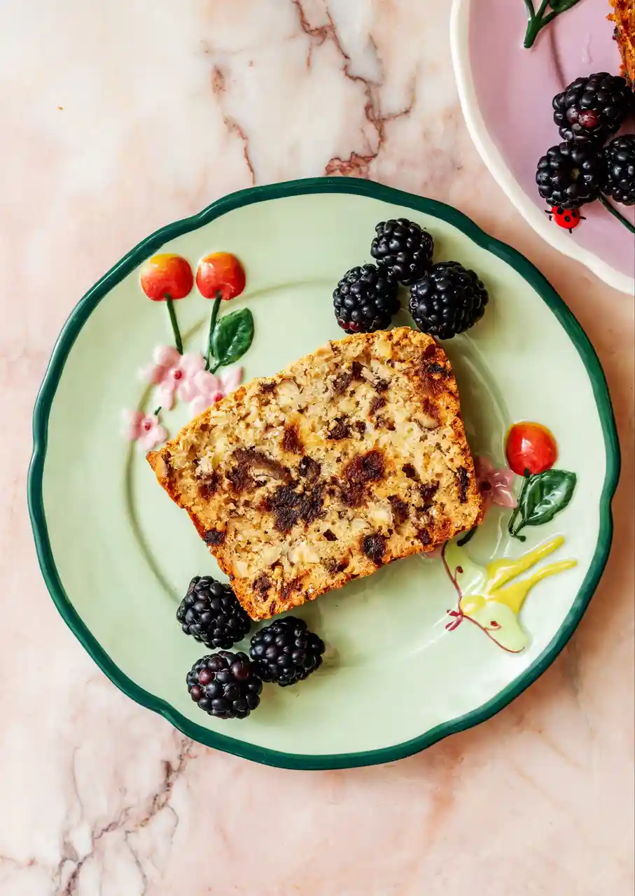 A slice of fruit bread with visible raisins and nuts sits on a light green plate with a dark green rim, decorated with…