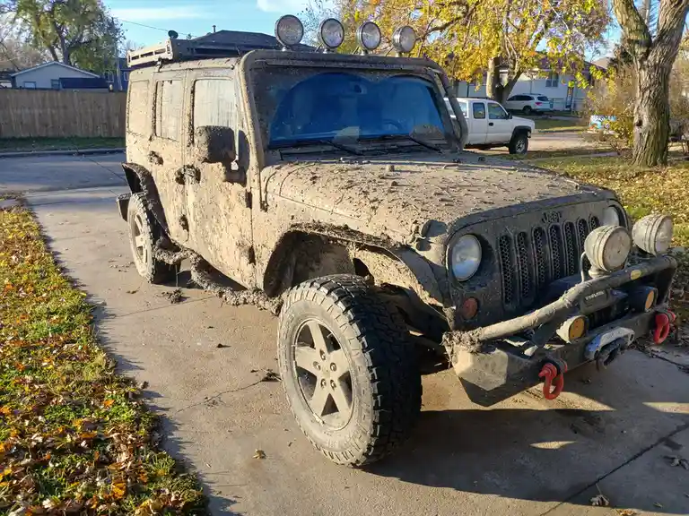 A heavily mud-splattered black Jeep Wrangler, equipped with roof-mounted lights and a front bumper with red recovery hooks…