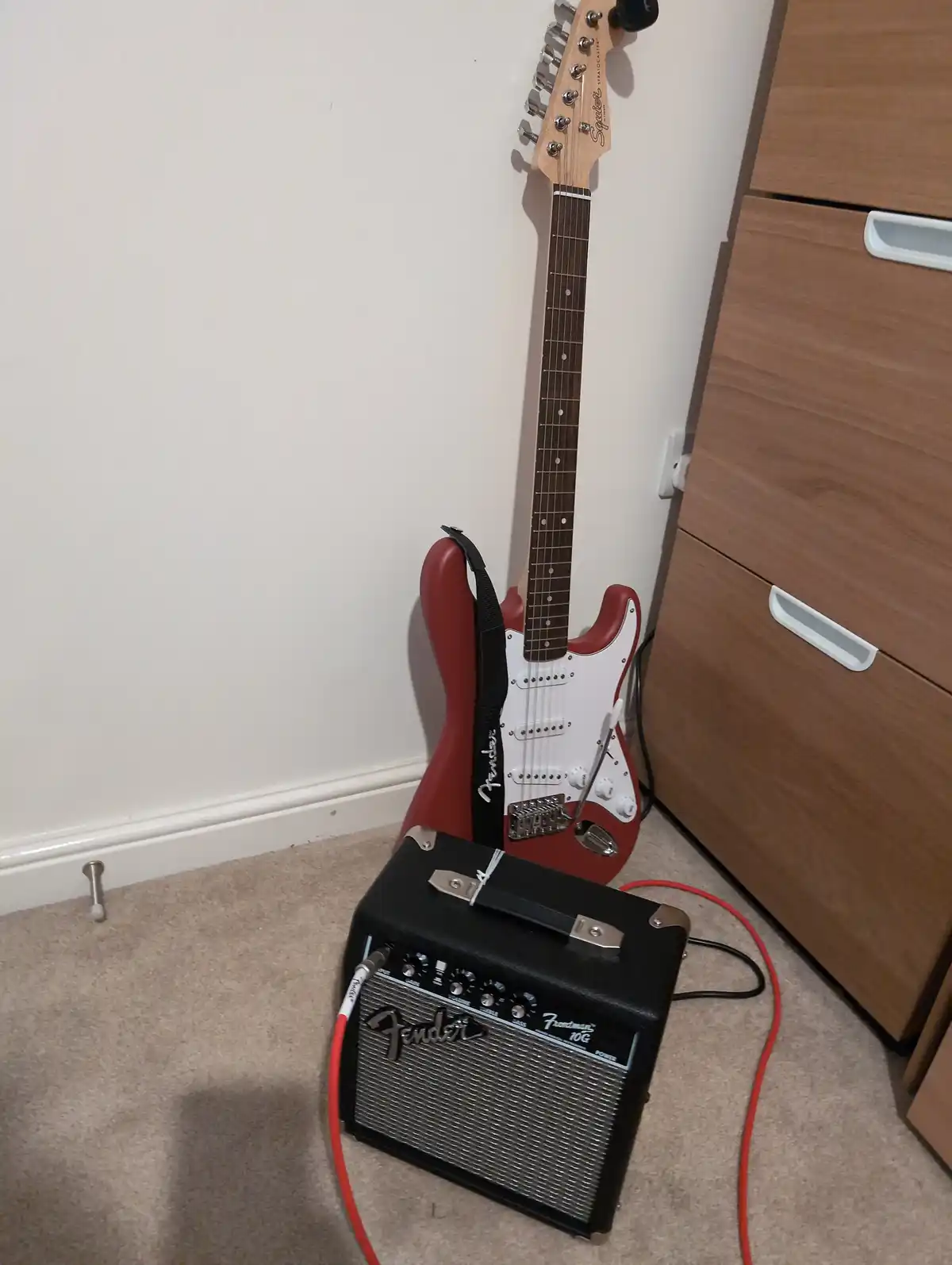 A red and white Fender Squier Stratocaster electric guitar leans against a white wall next to a wooden cabinet, with a black…