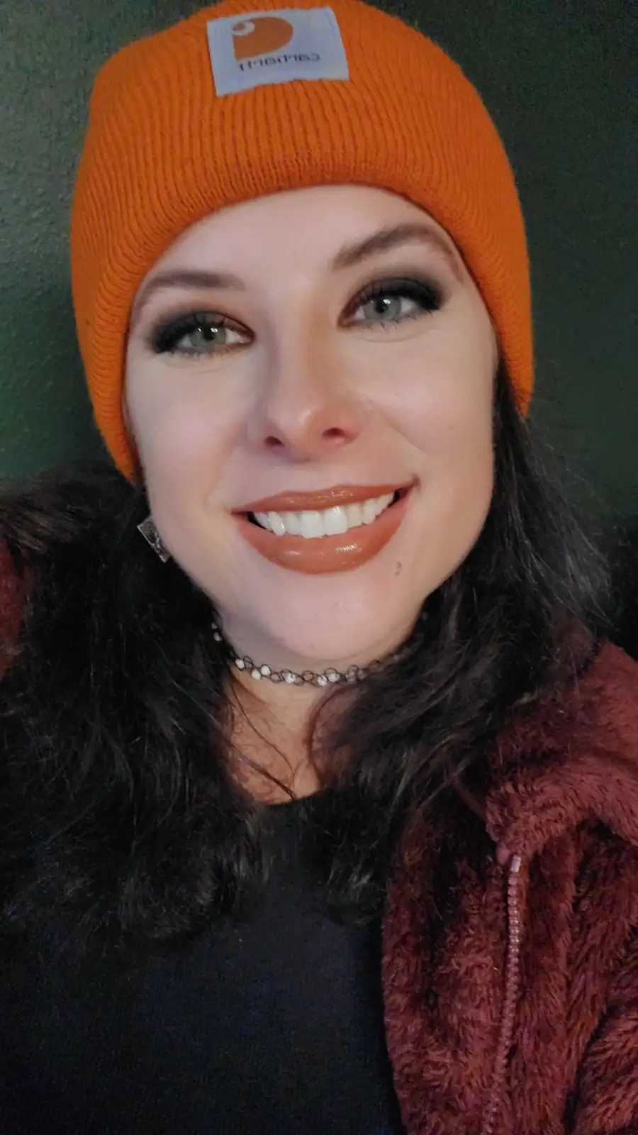 A woman with dark hair and makeup smiles while wearing a bright orange Carhartt beanie, a dark top, and a fuzzy…