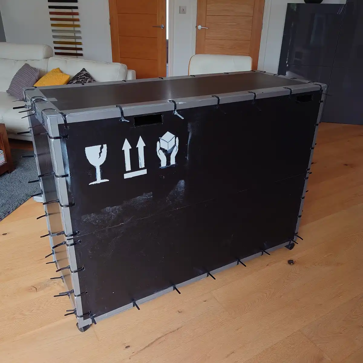 A large, black, rectangular shipping crate with silver metal edges and black straps is sitting upright on a light wood floor…