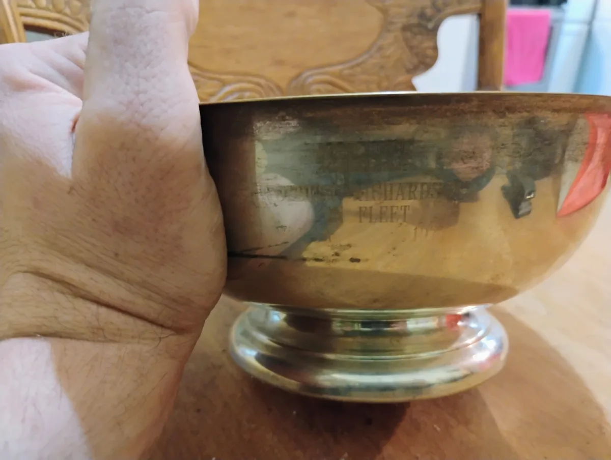 A close-up photo shows a hand holding a tarnished silver bowl with engraved text that reads 'DENNIS RICHARDS IN FLEET'. The…