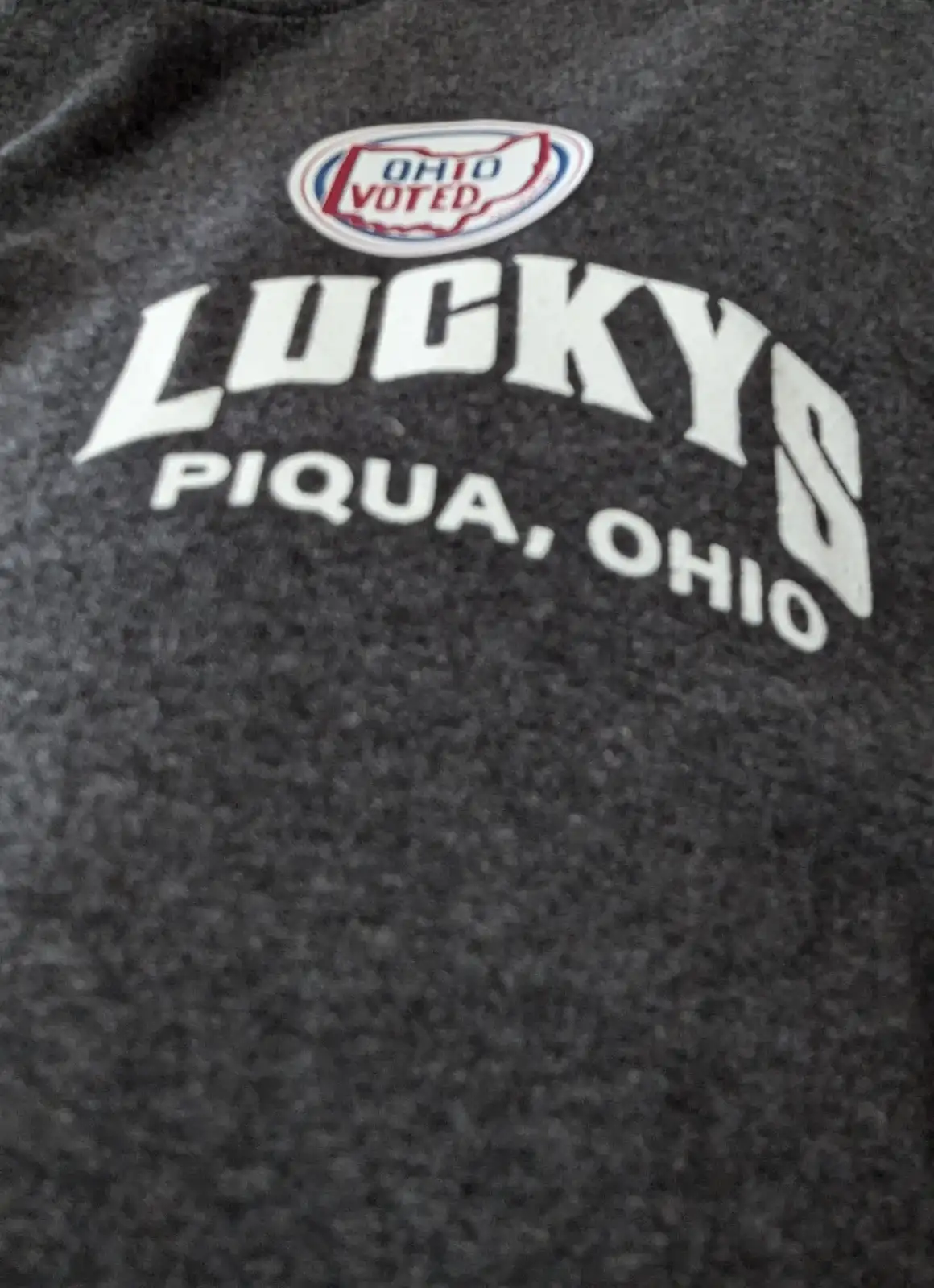 A close-up of a dark gray t-shirt with white text reading 'LUCKY'S PIQUA, OHIO' and a small oval logo above it that says…