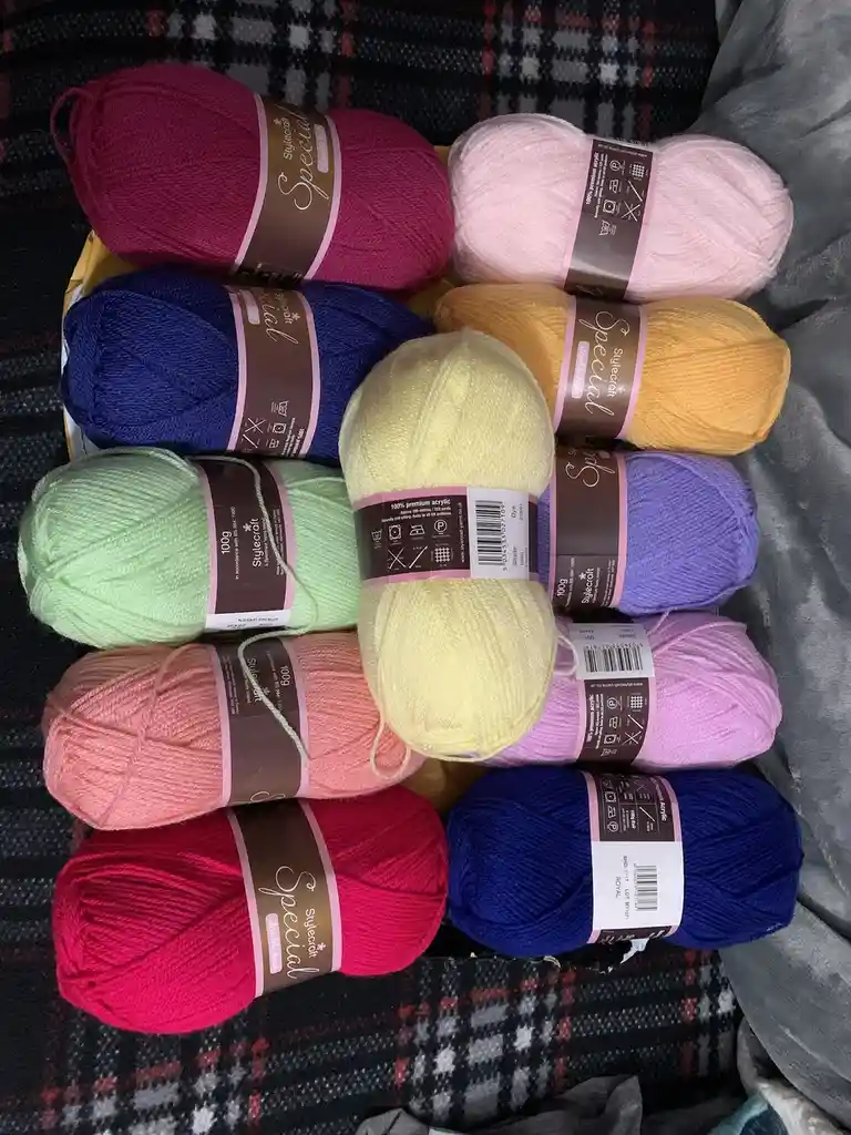 An overhead shot shows ten skeins of Stylecraft Special double knit yarn arranged in a grid on a plaid blanket, featuring a…