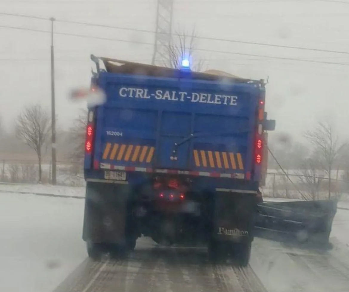 A blue snowplow truck is seen from behind on a snowy road, its rear has white text reading 'CTRL-SALT-DELETE'. The truck has…
