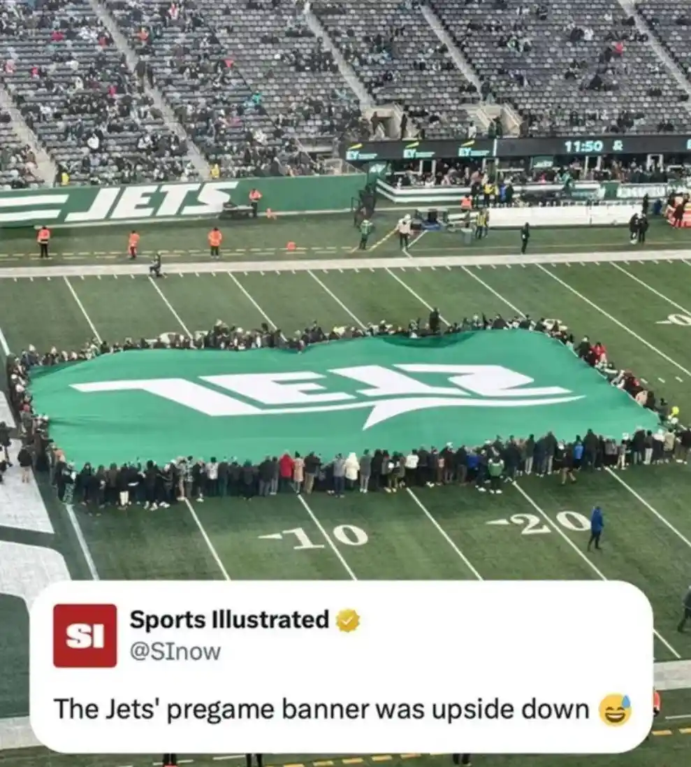 A large green banner with the New York Jets logo is laid out on a football field, surrounded by people holding it up. The…