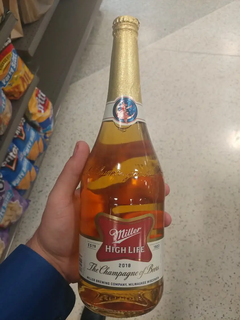 A hand holds a Miller High Life beer bottle in a store aisle, featuring a gold foil neck and label that reads 'The Champagne…