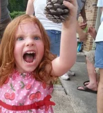 A young girl with red hair and a pink cherry-patterned dress is holding up a pinecone with an excited, wide-mouthed…