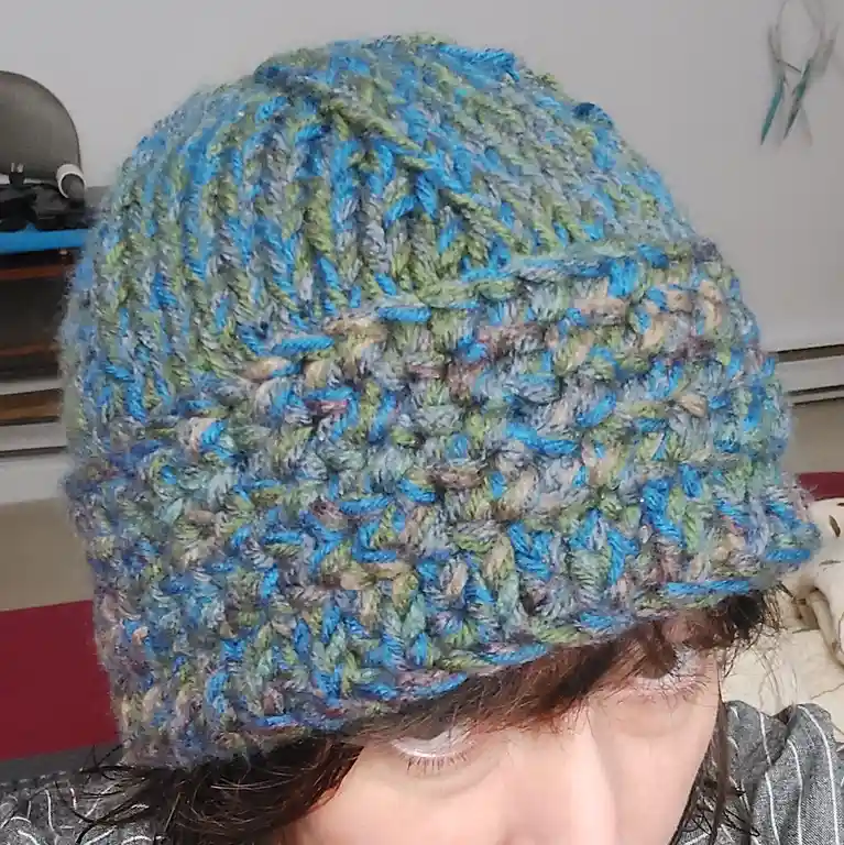 A close-up photo shows a person wearing a colorful, textured crocheted hat made of blue, green, and gray yarn. The hat…