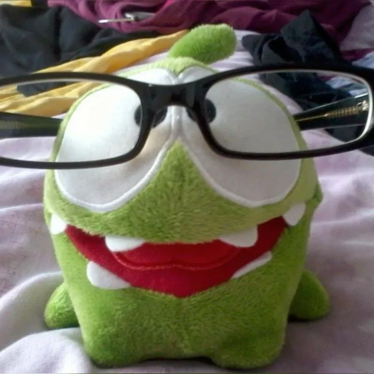 A green plush toy resembling the character Om Nom from the game 'Cut the Rope' has a pair of black-rimmed glasses pushed up…