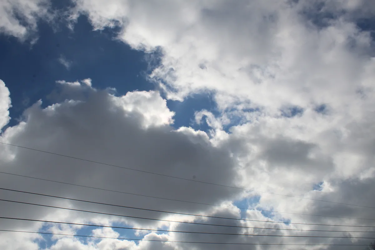 A photograph of a partly cloudy blue sky with dark grey clouds and several thin power lines crossing horizontally across the…