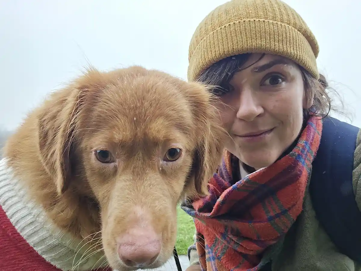 A person wearing a yellow beanie and plaid scarf poses with a dog in a sweater, both looking at the camera in a snowy…