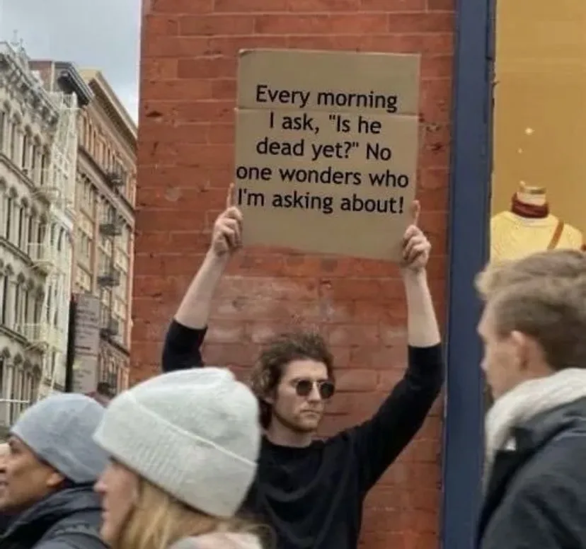 A man with curly hair and sunglasses holds up a cardboard sign with black text against a brick wall in a city street. The…
