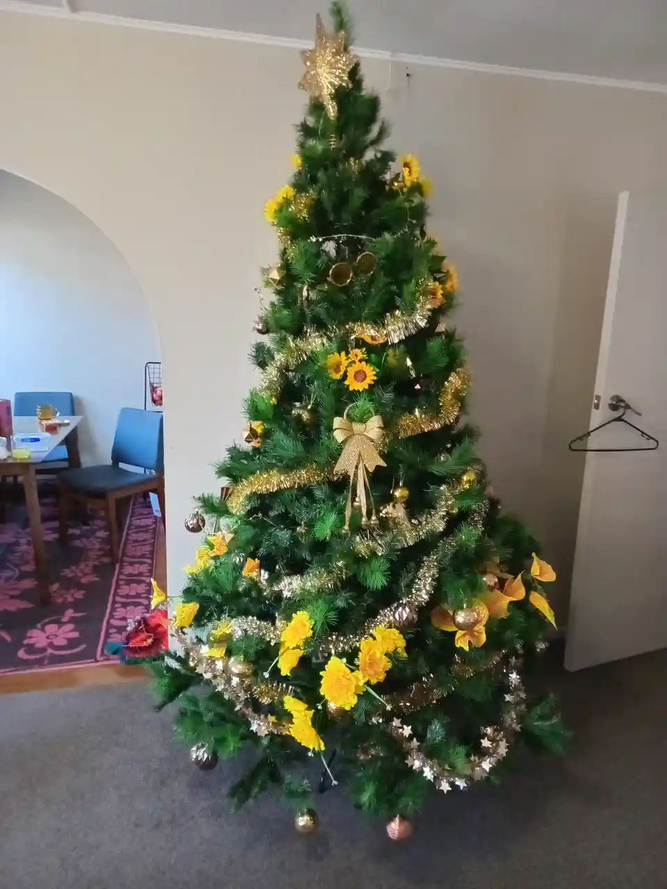 A decorated christmas tree adorned with gold tinsel, ornaments, and bright yellow sunflowers, standing in a room with a…