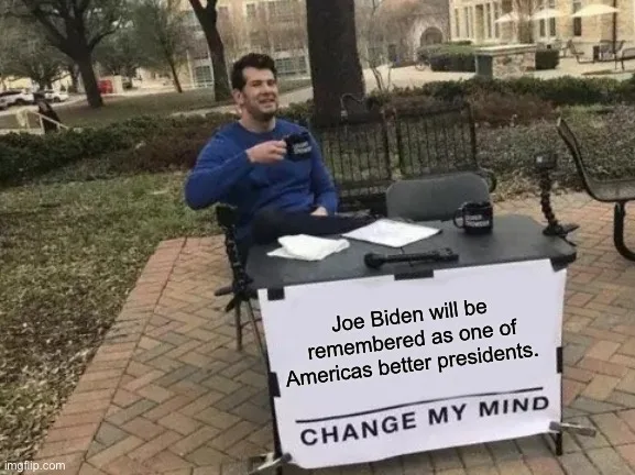 A man in a blue sweatshirt sits at a small outdoor table with a sign that reads 'Joe Biden will be remembered as one of…
