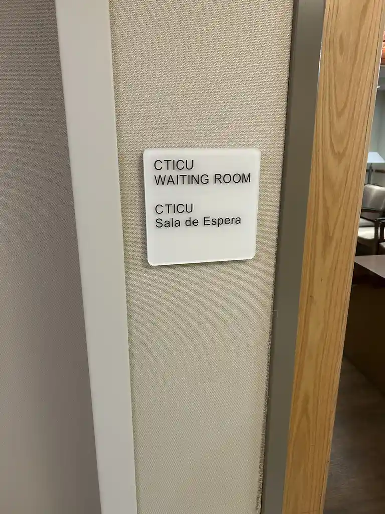 A close-up photo of a white rectangular sign on a beige fabric door frame, showing 'CTICU WAITING ROOM' in English and…