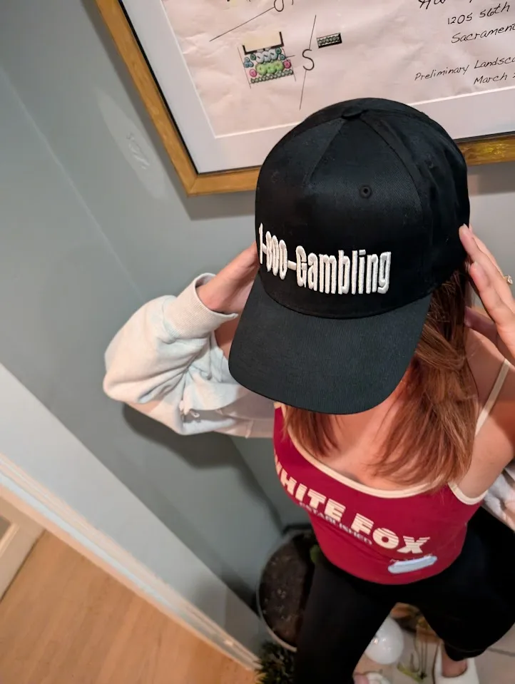 A person viewed from behind, wearing a black baseball cap with the text '1-900-Gambling' in white lettering, adjusts the cap…