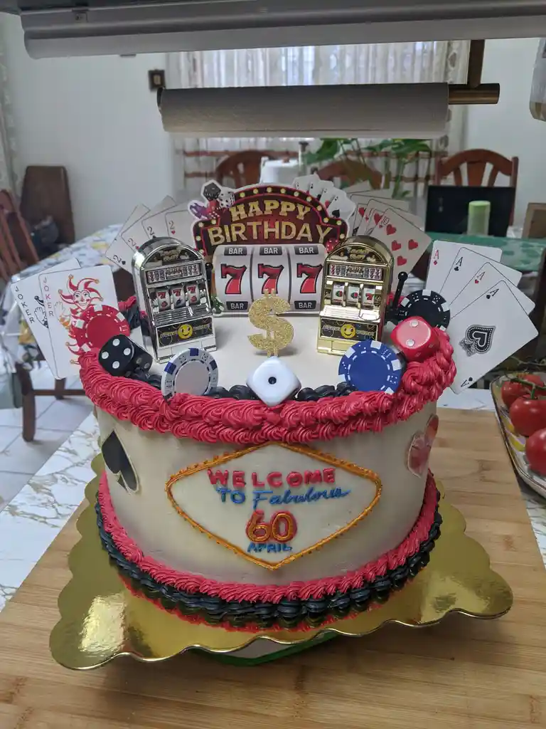 A birthday cake decorated with casino-themed items including miniature slot machines, playing cards, dice, and poker chips…