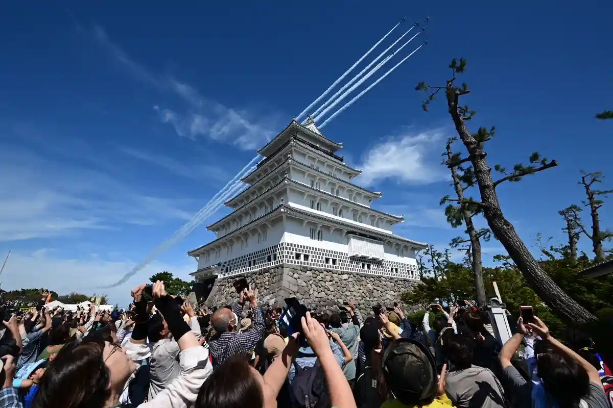 A crowd of people with phones raised watches jets flying in formation, leaving white smoke trails, against a clear blue sky…