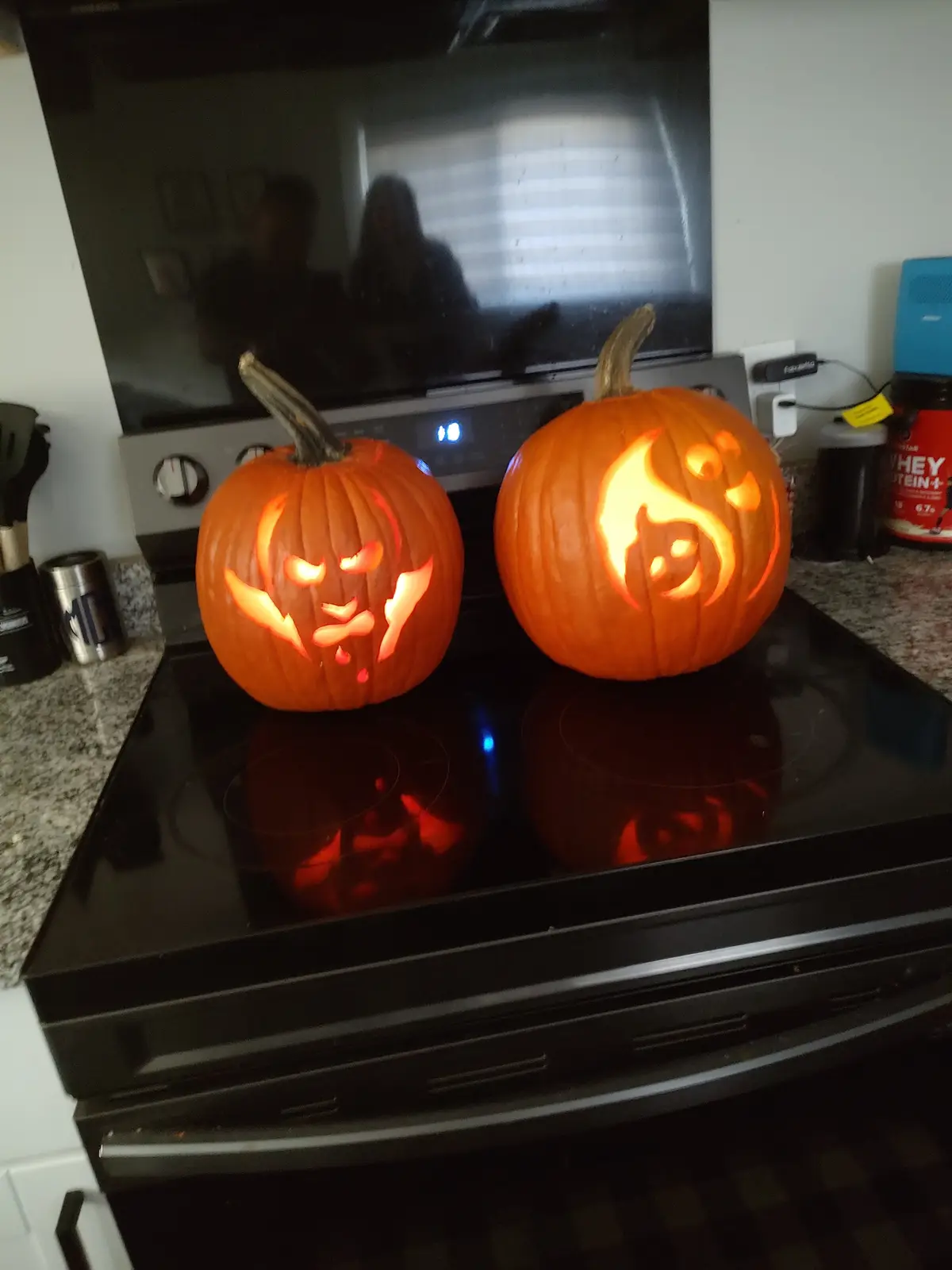 Two carved Halloween pumpkins sit on a black electric stove, glowing from within. The pumpkin on the left has a menacing…