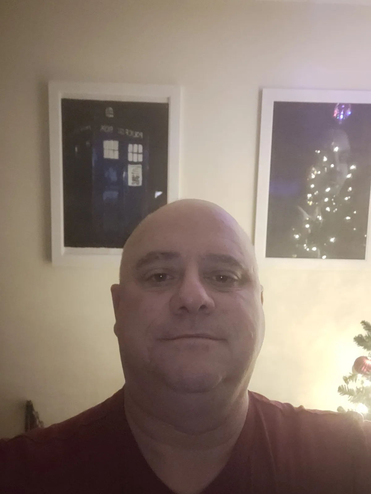 A bald man in a red shirt takes a selfie indoors, with two framed pictures on the wall behind him: one of a blue police box…