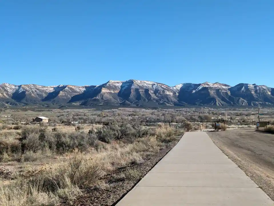 A wide-angle photo shows a paved sidewalk leading toward distant snow-capped mountains under a clear blue sky, with dry…