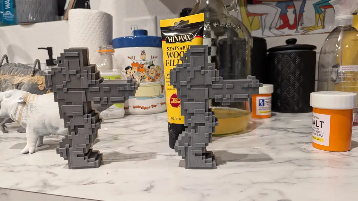 Two gray, pixelated, 3D-printed gun-shaped objects stand on a kitchen counter next to a bottle of Minwax wood filler and a…
