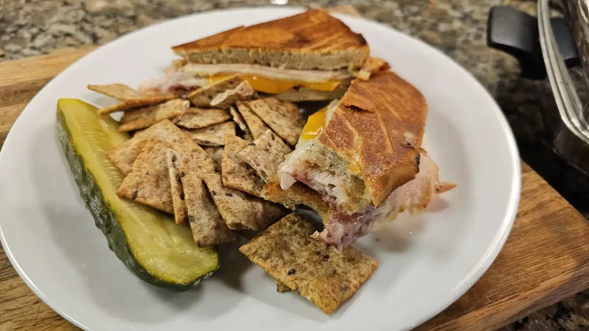 A white plate holds a toasted sandwich cut in half, revealing melted cheese and meat, alongside a large pickle spear and a…