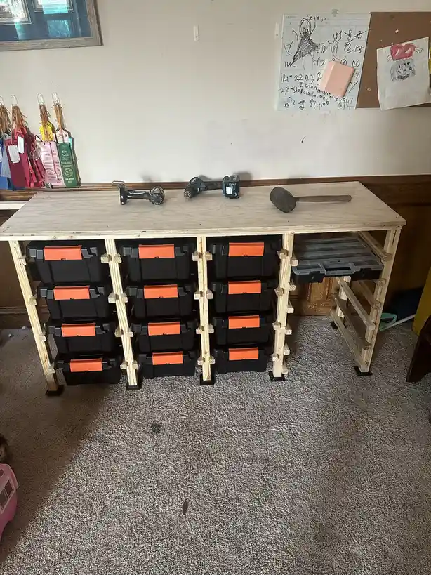A homemade wooden workbench with multiple black and orange plastic storage bins stacked underneath, topped with a drill…