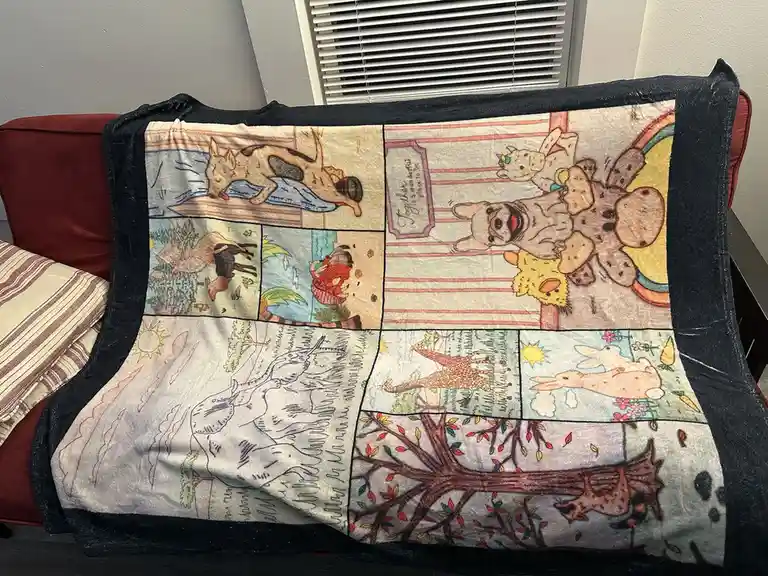 A cozy fleece blanket with a black border is draped over a red couch, featuring a patchwork of illustrated scenes with…