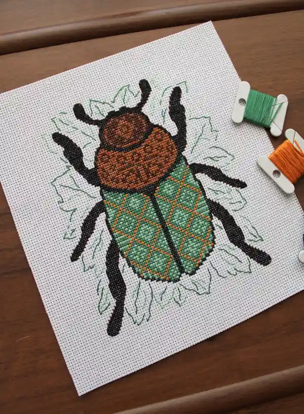 A partially stitched cross-stitch pattern of a beetle with a brown head, green body with orange grid lines, and black legs…