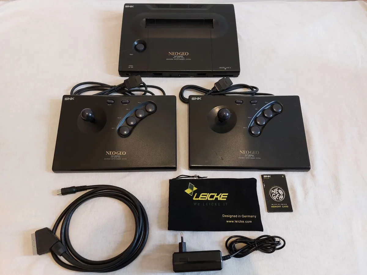 A complete SNK Neo-Geo AES console set laid out on a white surface, including the main unit, two black arcade-style…