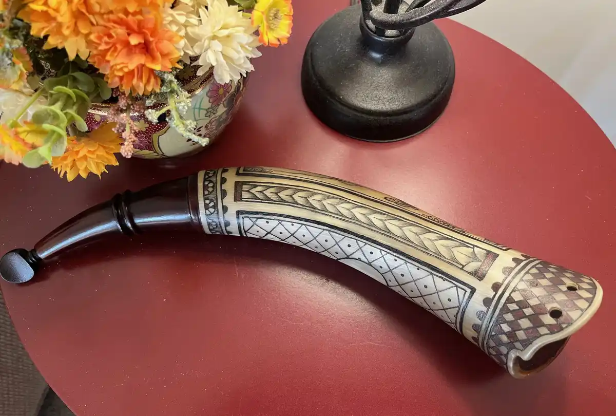 a curved, intricately decorated horn with geometric and leaf-like patterns rests on a red table, next to a vase of orange…