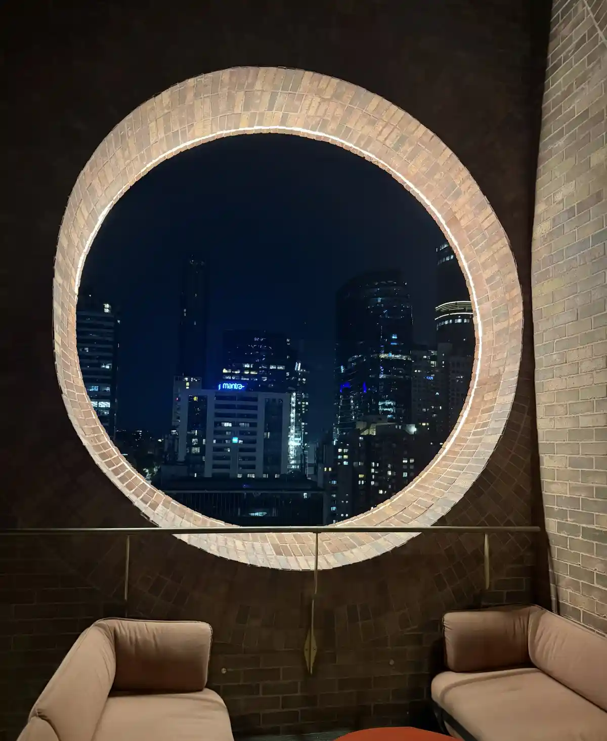 A nighttime photo shows a large circular brick window framing a city skyline with lit skyscrapers, including one with a…