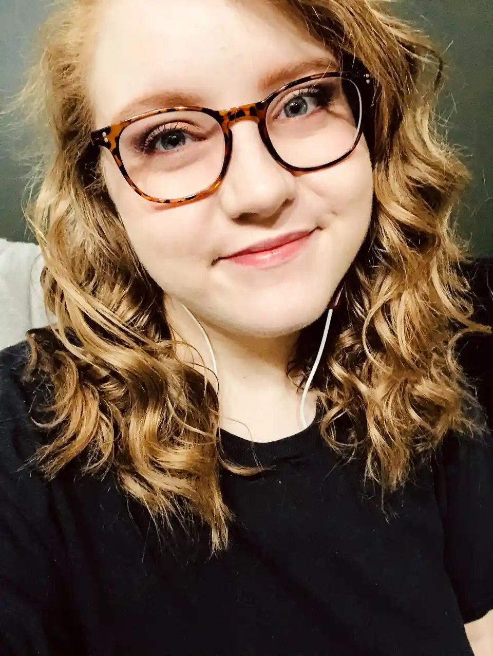 A close-up selfie of a young woman with curly blonde hair, wearing tortoiseshell glasses and white earbuds, smiling slightly…